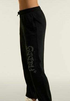 Pantalones deportivos negros con cintura elástica, bolsillos laterales y diseño de texto con cristales en la pierna, usados con zapatillas blancas.