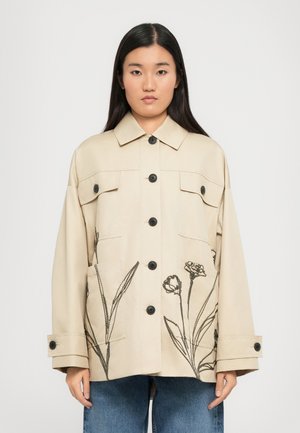 Veste beige avec un devant à boutons, deux poches poitrine et des broderies florales noires sur la partie inférieure. Design uni avec une coupe décontractée.
