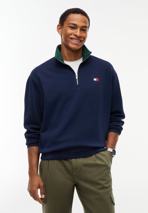 Uomo sorridente che indossa una felpa blu navy con zip al quarto, collo verde, maglietta bianca, pantaloni cargo color oliva e un orologio da polso argento.