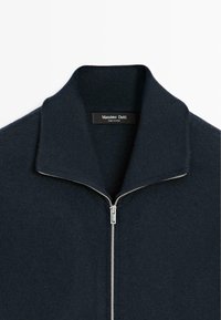 Námořnický svetr na zip s ribovaným límcem, který má stříbrný zip a štítek "Massimo Dutti." Měkká pletená textura, minimalistický design.