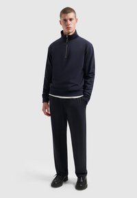 Marineblauwe sweatshirt met rits en een getailleerd silhouet, zachte textuur en geribbelde manchetten, gecombineerd met bijpassende losse broek en zwarte schoenen.