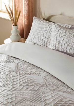 Weißes Bettüberwurf und Kissenbezüge mit strukturierten geometrischen Mustern auf einem Bett neben einem hölzernen Nachttisch mit einer Vase mit getrocknetem Gras.