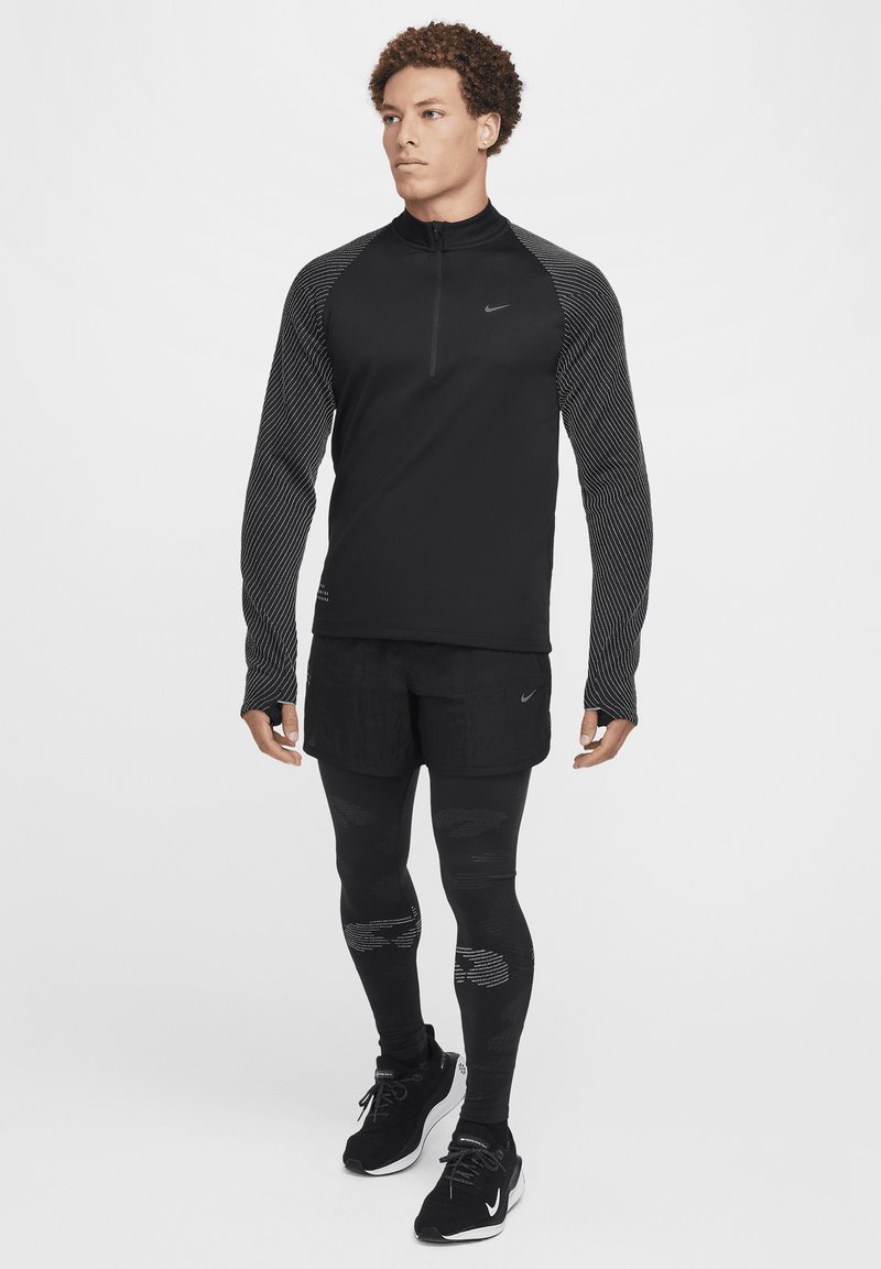 Haut noir à manches longues Nike avec fermeture éclair quart et manches texturées, associé à un short noir et des leggings à motifs, porté avec des baskets noires.
