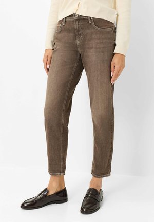 Person iført brune jeans med lige ben, en cremefarvet sweater og sorte læder loafers står foran en ensfarvet hvid baggrund.