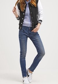 Varsityjack in zwart en wit, blauw gestreept overhemd, slim-fit blauwe jeans en witte sneakers met zwarte accenten. Casual outfit.