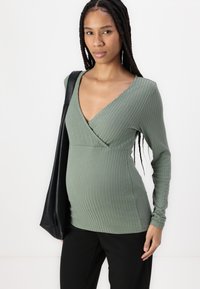 T-shirt long à manches en vert olive côtelé avec un décolleté en V profond et une coupe ajustée. Le mannequin tient un sac noir, associé à un pantalon noir.