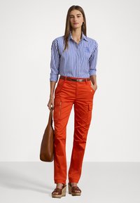 Gestreepte blauw-witte shirt in combinatie met fel oranje cargobroek, voorzien van ritsen en zakken. Bruine schoudertas en platformsandalen.