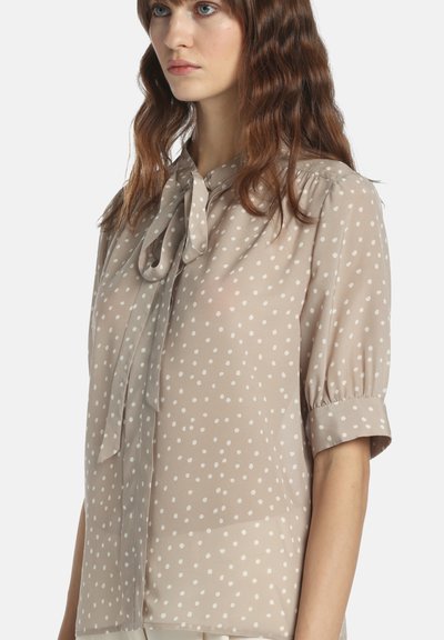 Prosojna bež bluza z belimi pikami, kratkimi puhastimi rokavi in zavezovanjem na izrezu vratu. Lahka tkanina in mehka tekstura.