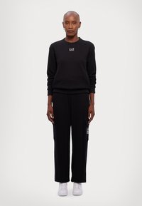 Zwarte sweatshirt met een logo op de borst, gecombineerd met zwarte broeken met zijzakken. Onder zijn witte sneakers.