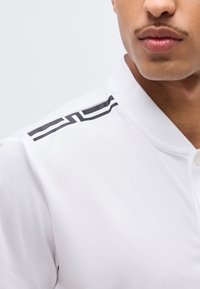 Κλείσιμο εικόνας ενός ατόμου που φοράει λευκό πουκάμισο με γιακά και μαύρη γεωμετρική λωρίδα στον ώμο, εμφανίζοντας τα χείλη και το πηγούνι.