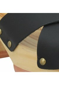 Sangle en cuir noir avec des clous en laiton attachée à une base en bois clair, présentant une forme courbée et une texture lisse.