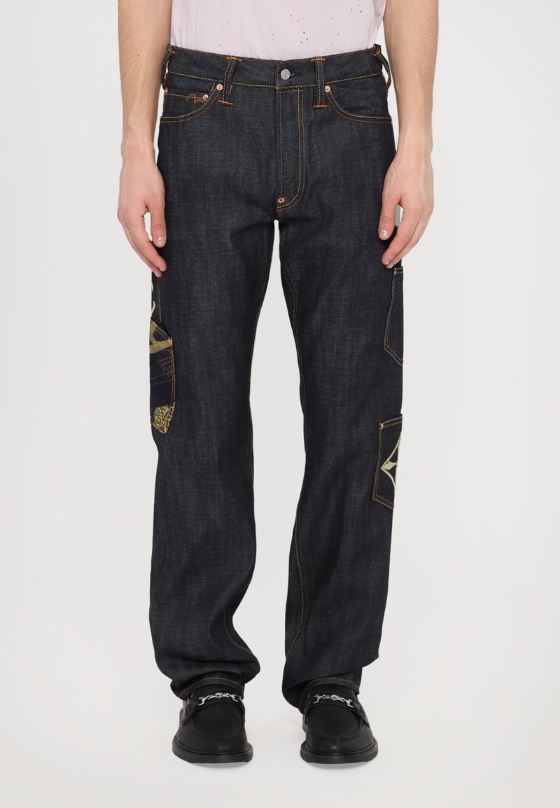 Pantalon en denim bleu foncé avec des coutures orange contrastantes, accents de patchwork uniques sur les poches, coupe décontractée et design cinq poches.