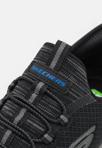 Black Skechers sneaker featuring a textured mesh upper, grey and black elastic laces, and a contrasting blue logo tag.