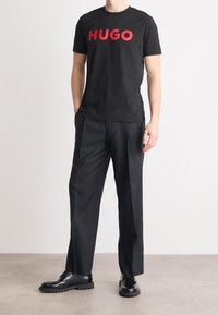 Schwarzes T-Shirt mit auffälligem roten "HUGO"-Schriftzug, kombiniert mit schwarzen maßgeschneiderten Hosen und schwarzen Schnürschuhen, vor einem neutralen Hintergrund.