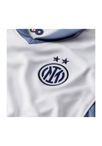 Maillot de sport blanc avec un col bleu ; il présente un logo brodé en marine avec deux étoiles au-dessus, fabriqué en tissu lisse et respirant.