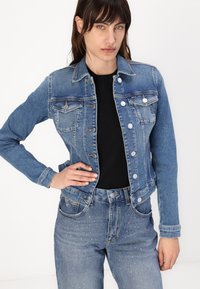 Sininen farkkutakki, jossa on kaulus, etutaskut ja hopeanväriset napit. Päällä musta paita ja kevyet vaaleansiniset farkut, joissa on kimallusta.