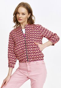 Gemusterte Bomberjacke in pink- und cremefarbenem geometrischen Design, mit Reißverschluss vorne, elastischen Bündchen und Bund. Kombiniert mit hellpinken Hosen.