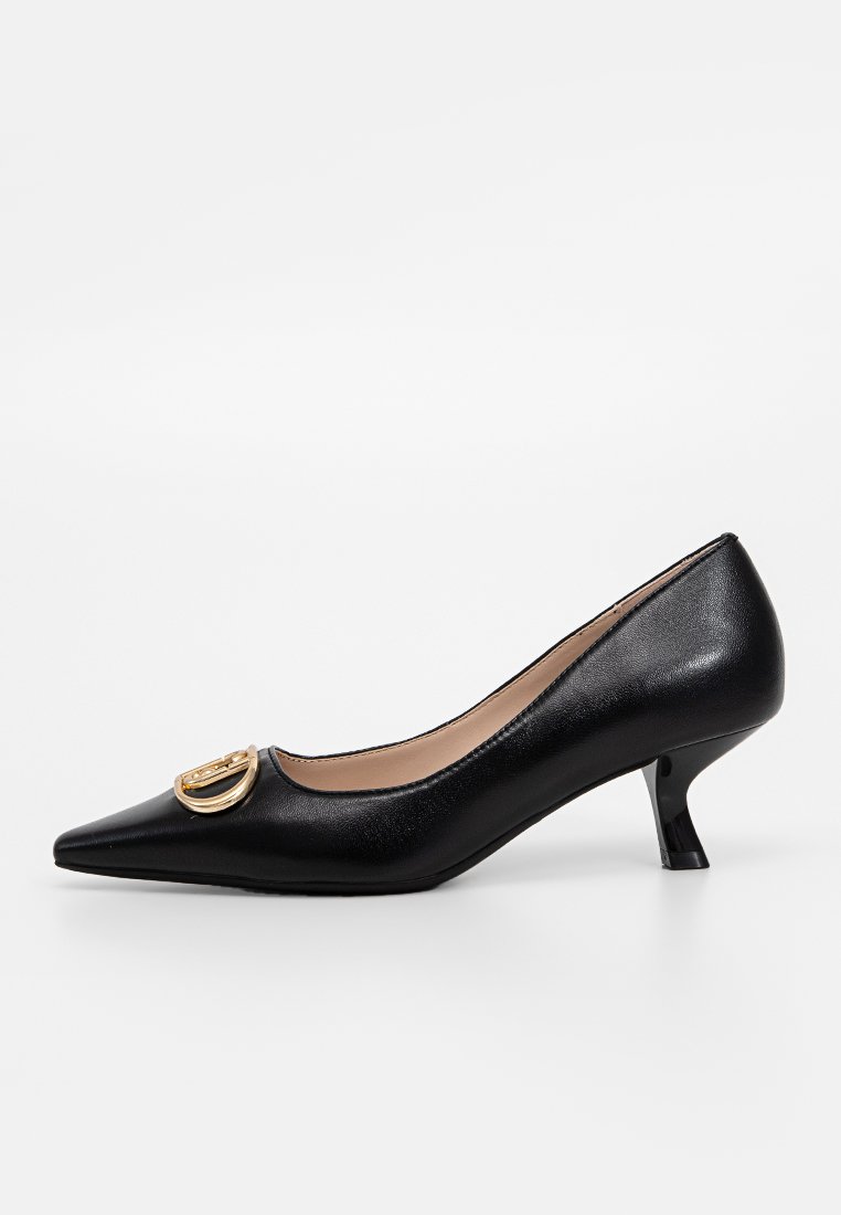 Zapato de tacón negro de cuero con punta afilada y un tacón pequeño y curvado. Presenta un acento circular dorado en la parte superior y una textura suave.