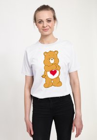 Vit bomullströja med en tecknad björn som har ett hjärta på magen. Björnen är brun med ett ljust ansikte och håller sina tassar samman.