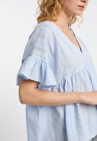 Lichtblauwe blouse met een diepe V-hals, korte, gerimpelde mouwen, verticale strepen en kantdetails. Gelaagd stof met een losse pasvorm.