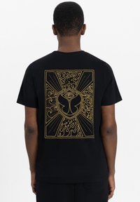 Zwarte t-shirt met een groot gouden grafisch ontwerp op de achterkant, met krullen, vlammen en een centraal embleem dat op een vlinder lijkt.