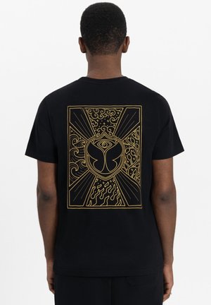 T-shirt noir avec un grand motif doré au dos, présentant des tourbillons, des flammes et un emblème central ressemblant à un papillon.