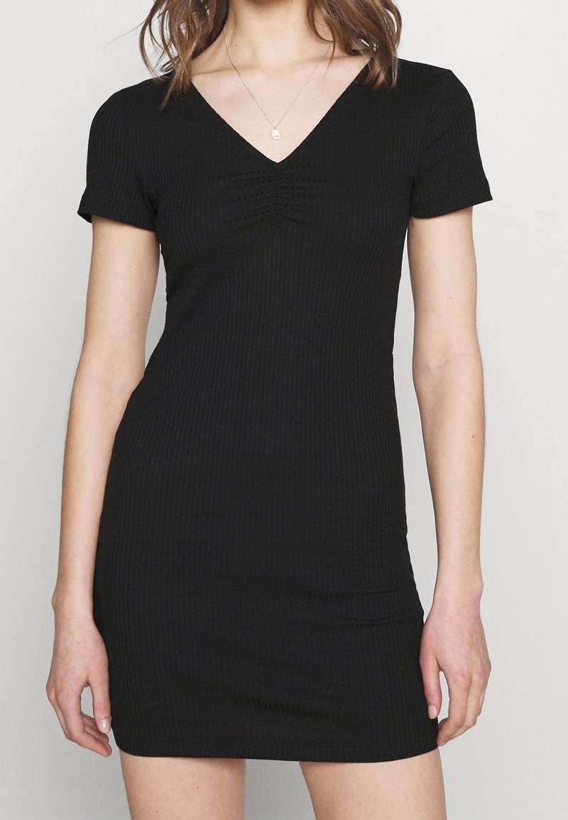 Kvinna iklädd en kort svart ribbad klänning med korta ärmar och V-ringning, accessoriserad med ett litet runt hängande smycke.