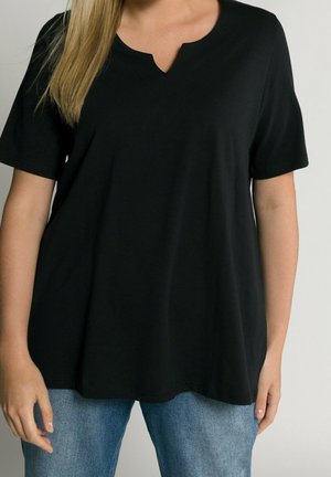 Frau trägt ein schlichtes, lockeres schwarzes T-Shirt mit V-Ausschnitt und kurzen Ärmeln sowie blaue Jeans, steht vor neutralem Hintergrund.
