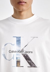 Bílá bavlněná tričko s velkým grafickým potiskem písmen "cK" v modré a šedé barvě, s textem "Calvin Klein Jeans" v černé barvě pod ním.