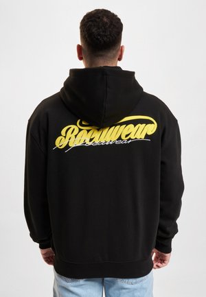 Mann mit kurzen lockigen Haaren, der einen schwarzen Hoodie mit gelbem und weißem "Rocawear" Text auf dem Rücken trägt, hellblaue Jeans, mit dem Rücken zur Kamera auf weißem Hintergrund.