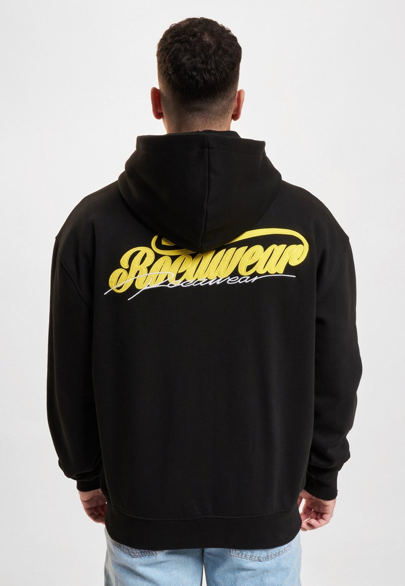 Homme aux cheveux courts et bouclés portant un sweat à capuche noir avec un texte "Rocawear" jaune et blanc dans le dos, jean bleu clair, vu de dos sur fond blanc.