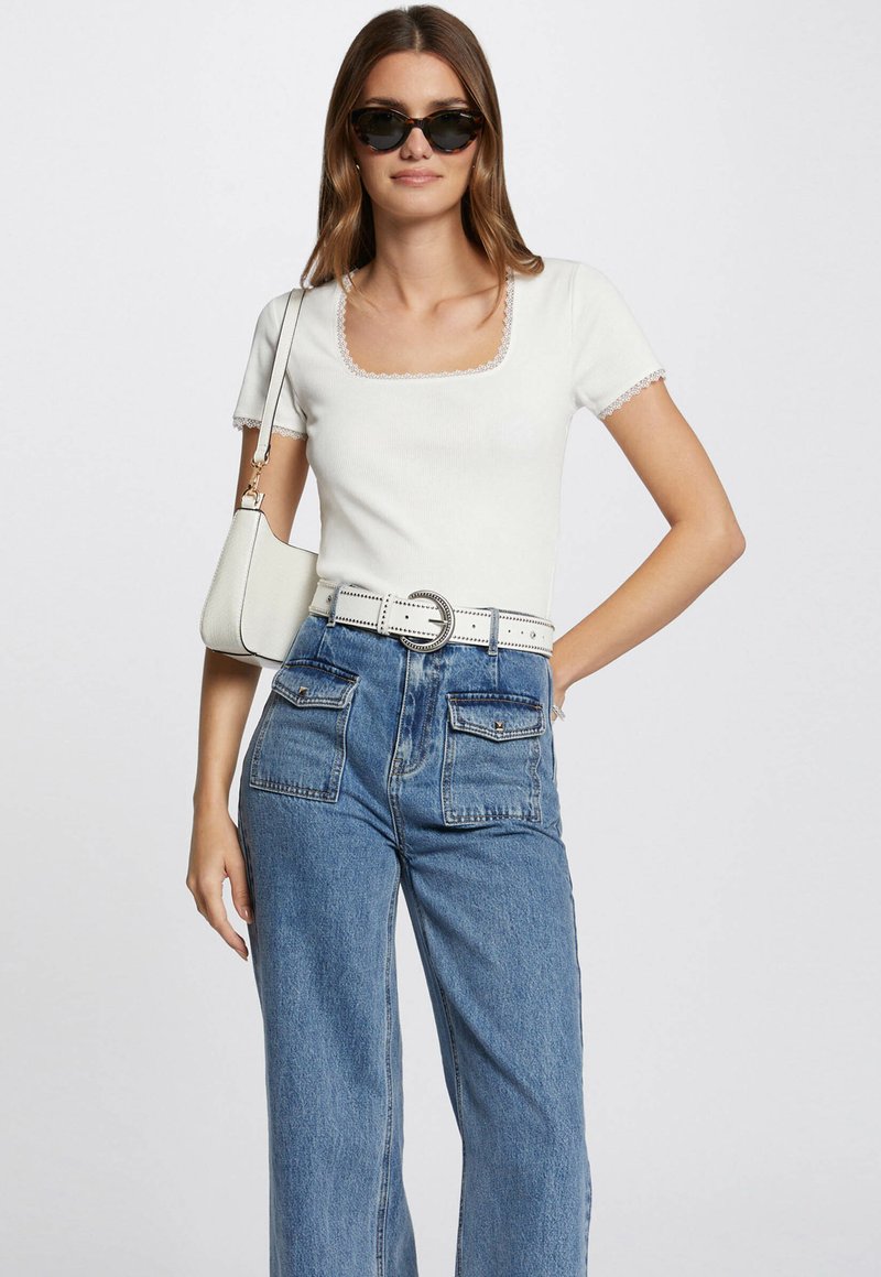Un top blanc à manches courtes avec un décolleté carrée et une dentelle, associé à un jean en denim bleu taille haute avec de grandes poches avant. Tenant un sac crème.