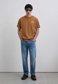 Camiseta a rayas marrón con logo, combinada con jeans de mezclilla azul claro y zapatos marrones oscuros. El modelo lleva gafas de sol negras. Fondo simple.