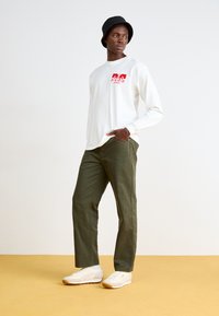 Une chemise blanche à manches longues avec un motif rouge, un pantalon vert olive et des baskets de couleur claire. Le modèle porte un chapeau seau noir. Fond simple.