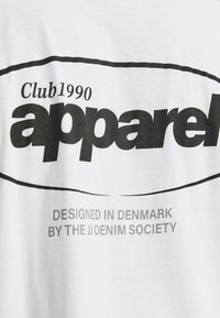 Camiseta blanca de algodón con un texto negro en negrita que dice "Club1990 apparel", con una línea curva y un texto más pequeño abajo que indica "DISEÑADO EN DINAMARCA POR LA JJ DENIM SOCIETY".