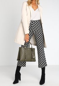 Olivengrønt håndtaske med præget geometrisk mønster, dobbelte top håndtag og sølvfarvet hardware, parret med stribede sort-hvide bukser.