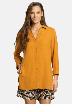 Camisa de botones amarilla mostaza con mangas largas, cuello y dos bolsillos laterales. Los pantalones cortos tienen un diseño abstracto en negro, blanco y naranja.