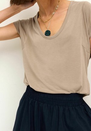 Vrouw draagt een beige korte mouwen V-hals top, gelaagde gouden kettinkjes met een donker groene hanger, en een zwarte rok met elastische taille.