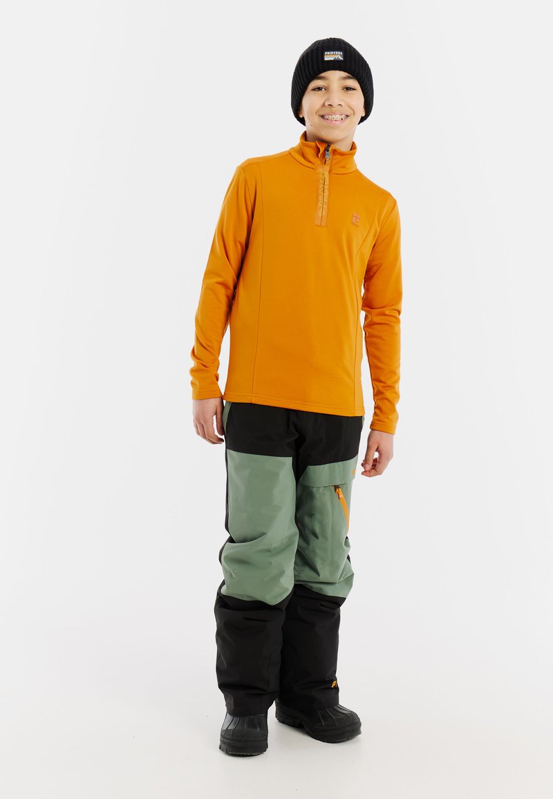 Pomarańczowy sweter z długim rękawem i zamkiem błyskawicznym w ćwierć długości, zestawiony z czarnymi i zielonymi spodniami narciarskimi z zamkami błyskawicznymi w kieszeniach i wzmocnieniami na kolanach. Czarna czapka na głowie.