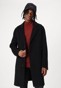 Sort uldovertøj med en klapkrave, enkeltknaplukning og sidelommer, båret over en rød turtleneck sweater.