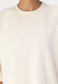 Lys beige genser med rund hals som har ribbestrikket krage og korte raglanermer. Brodert merke tekst i en subtil nyanse på forsiden.