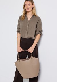 Beige Leder-Shopper mit strukturiertem Design, kurzen Griffen und glatter Textur. Das Model trägt ein hellbraunes Hemd mit Knöpfen und dunkle Hosen.