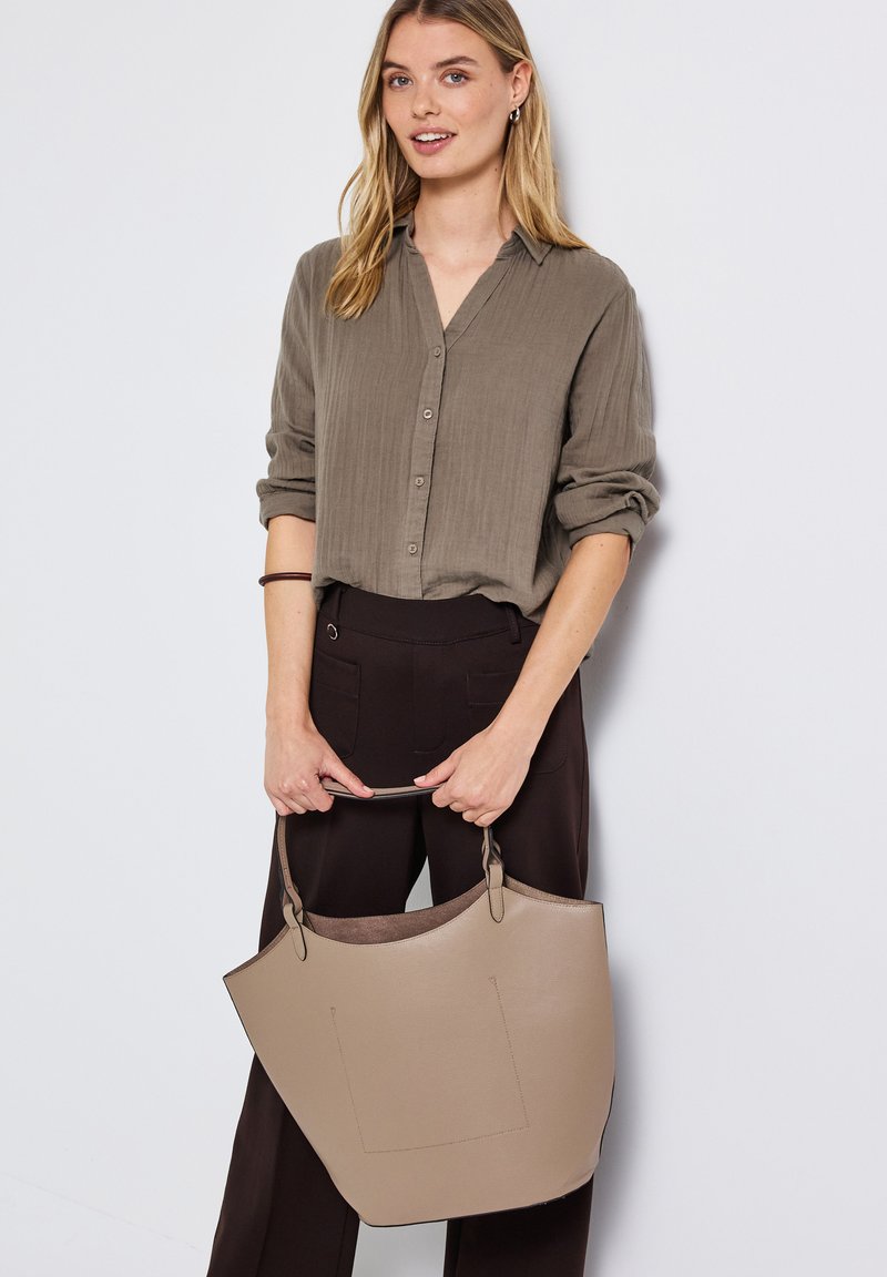 Beige Leder-Shopper mit strukturiertem Design, kurzen Griffen und glatter Textur. Das Model trägt ein hellbraunes Hemd mit Knöpfen und dunkle Hosen.
