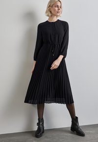 Schwarz plissiertes Kleid mit rundem Ausschnitt, Knopfverschluss und Kordelzug in der Taille. Getragen mit schwarzen Stiefeletten und durchsichtigen Strumpfhosen.