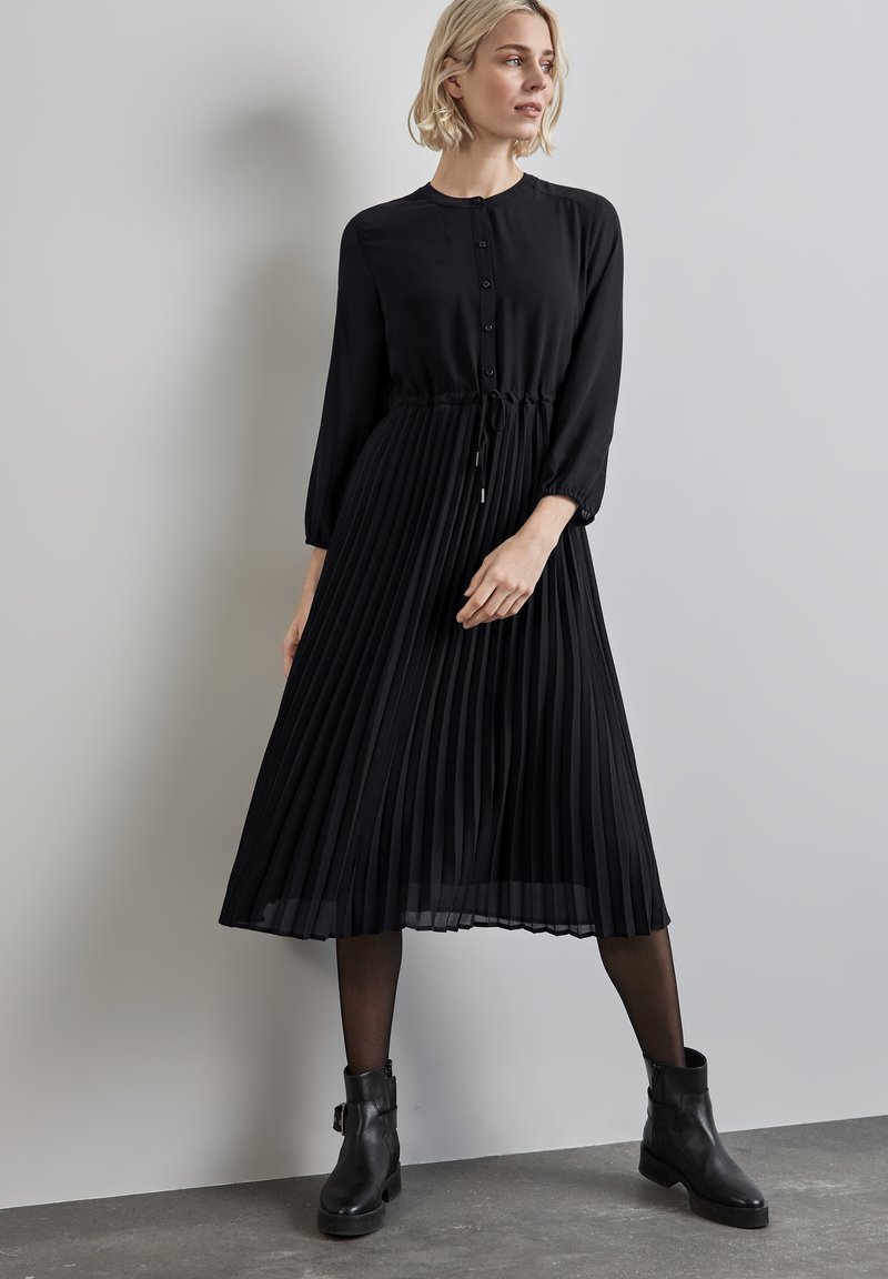 Schwarz plissiertes Kleid mit rundem Ausschnitt, Knopfverschluss und Kordelzug in der Taille. Getragen mit schwarzen Stiefeletten und durchsichtigen Strumpfhosen.