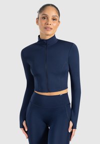 Marineblauwe cropjack met een hoge kraag en volle rits, lange mouwen en een gladde, rekbare stof. Gecombineerd met bijpassende high-waisted leggings.