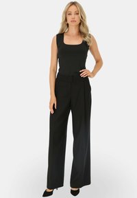 Czarny bezrękawnik i szerokie spodnie, charakteryzujące się strukturalnym wzornictwem i gładką teksturą, zestawione z czarnymi obcasami. Czyste tło.