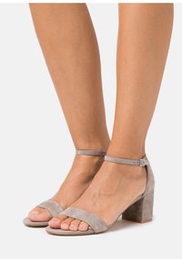 Sandales à talons en suède gris clair avec une large lanière au niveau des orteils et une lanière de cheville ajustable, présentant un talon bloc pour plus de stabilité.