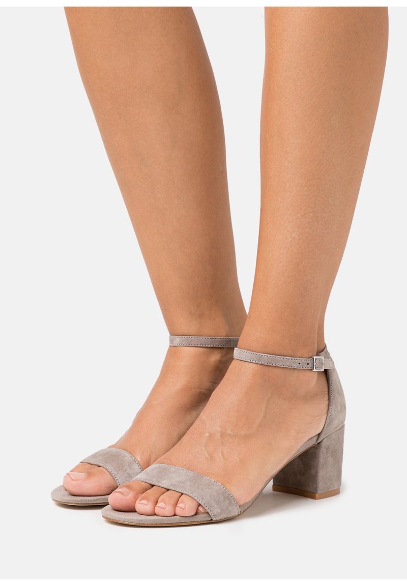 Sandales à talons en suède gris clair avec une large lanière au niveau des orteils et une lanière de cheville ajustable, présentant un talon bloc pour plus de stabilité.