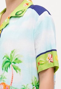 Camicia a maniche corte in tessuto azzurro chiaro con stampe tropicali di palme e fiori, colletto floreale verde e dettagli blu navy.